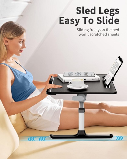 A young woman sitting comfortably on a bed, using an adjustable laptop table with a built-in storage drawer.