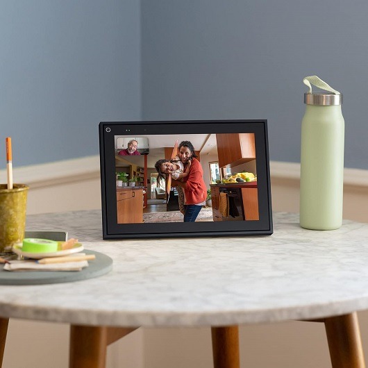 A black smart video calling device with a 10" touch screen display, sitting on a table in a living room setting.