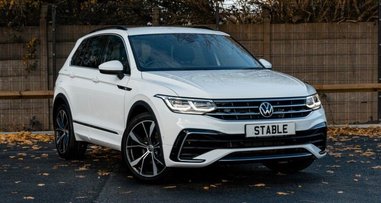  white 2021 Volkswagen Tiguan R-Line parked in front of a wooden fence. The Tiguan R-Line is a performance-oriented trim level of the popular Volkswagen SUV, offering a sporty look and powerful engine