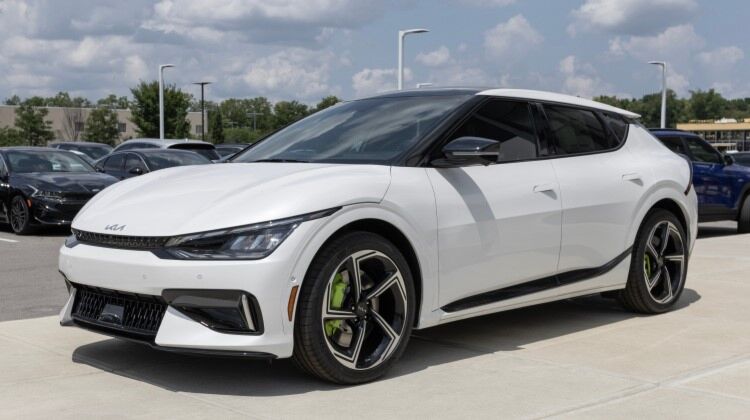 A white Kia EV6 GT electric car parked in a sunny parking lot. The Kia EV6 GT is a high-performance electric car with a curved sportback design and rear spoiler. It has a panoramic sunroof and black alloy wheels